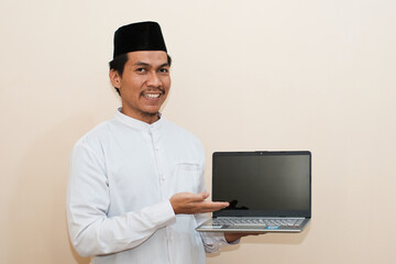 Smiling Southeast Asian Muslim man holding laptop showing blank screen. Cheerful person wearing muslim clothes presenting computer display