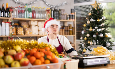 Male salesman in a Santa hat sits thoughtfully behind a counter with fruits and scales. Salesman in a supermarket alone celebrates the New Year