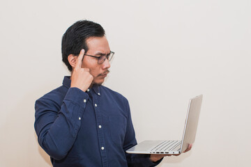 A Southeast Asian man wearing blue shirt with glasses holding laptop and thinking gesture, standing over isolated white background, Positive person