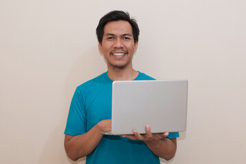 Smiling Southeast Asian man wearing green t-shirt casual clothes holding laptop, standing over isolated white background, Positive person