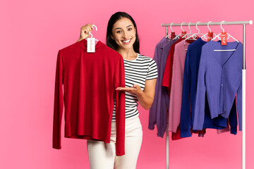 A young woman stands smiling in a colorful clothing store, holding a red sweater while showcasing...