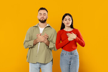 A young couple stands together, hands placed over their hearts, conveying deep affection. They are isolated against a bright yellow background, radiating warmth and intimacy.