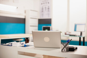 Digital laptop positioned on table in doctors office. Empty organized hospital room containing medical equipment and modern devices.