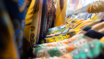 A close-up shot of colorful batik shirts hanging on wooden hangers in a market stall, with a selective focus on the front row and a blurred background
