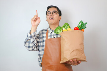 Asian Man with apron thinking gesture to grocery shopping bag with finger up with great new idea isolated on plain white