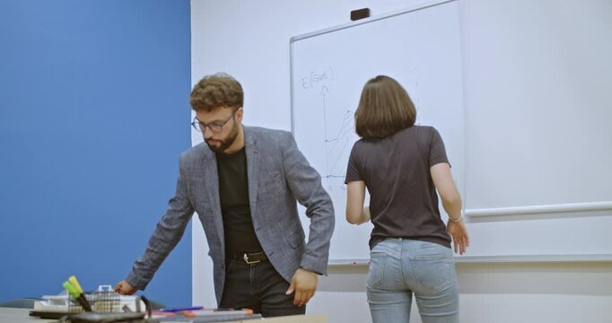 A female student explains a graph drawn on a whiteboard to a professor and group of students in a classroom. They're discussing a complex equation during a study session.