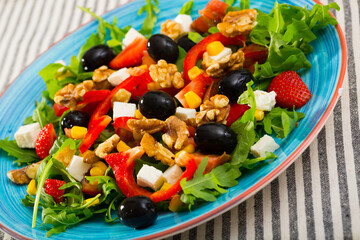 Fresh green arugula salad with vegetables, goat cheese and ripe strawberry decorated with black olives, walnuts and corn kernels