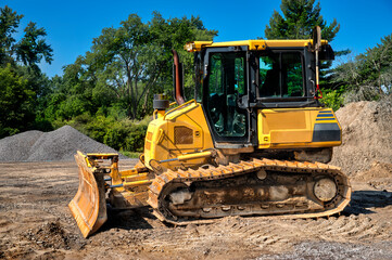 Yellow bulldozer with a front blade and caterpillar tracks. It’s situated in an area with piles of dirt or sand, surrounded by trees and a clear blue sky.