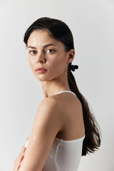 Obraz premium Portrait of a young woman with long dark hair and a ponytail, wearing a white sleeveless top, against a minimalistic light grey background, conveying beauty and simplicity