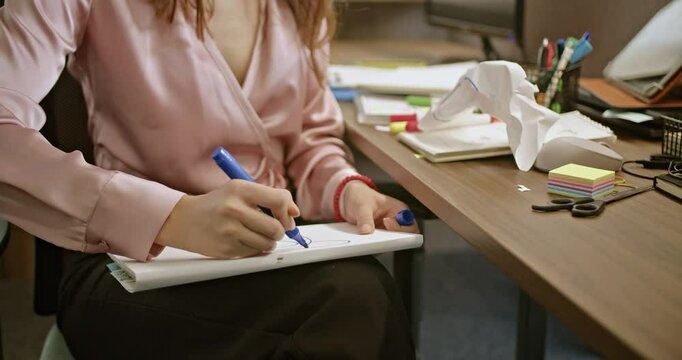 Woman doodling on paper at a messy desk in an office. She wears a pink shirt and black trousers, sitting and drawing circles. Office supplies and post-it notes visible.