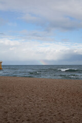 A sandy beach in Gangneung faces gentle waves, with a yellow beacon standing offshore. A faint rainbow arcs in the cloudy sky, adding charm to the serene seascape.