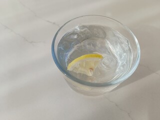 A clear glass of ice water with a slice of lemon, resting on a marble surface under soft sunlight.