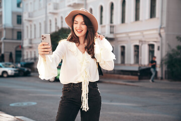 Portrait of young beautiful smiling hipster woman in trendy summer white blouse and jeans. Carefree woman posing in the street in sunny day. Positive model outdoors. Cheerful and happy, take selfie