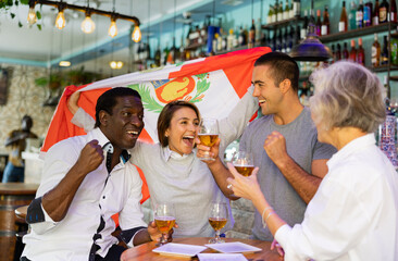 Obraz premium Happy sport fans holding flag of Peru, celebrating victory of national team, drinking alcoholic drinks in beer pub