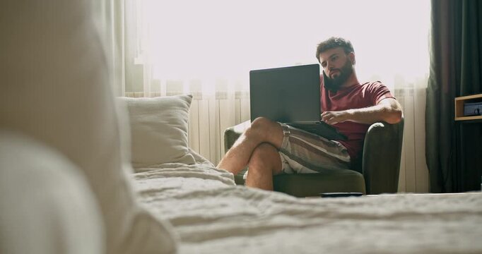 Young man working from home. He's sitting in his bedroom, wearing casual clothes and using a laptop. The room is bright and airy.