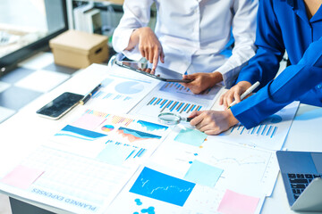 Two Asian business woman working and talking at a table with documents, a graph, and a digital tablet. They are focused on analysis, business planning, and collaborative discussions