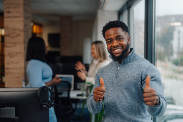 Smiling businessman gesturing thumbs up in modern office with coworkers clapping