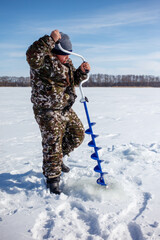 A man in camouflage clothing is standing in the snow with a blue ice auger