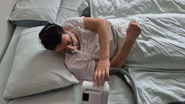 A man in his mid-thirties with sleep apnea sleeps using a nasal mask and CPAP device in a bed in his house