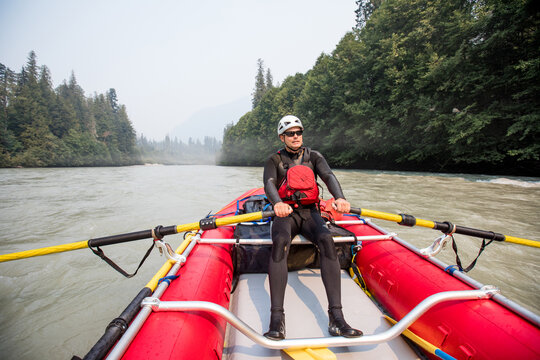 Man rowing oar frame raft on scenic river, shore lined with trees.