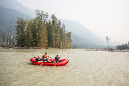 Two men paddling on red raft boat, forest fire smoke.