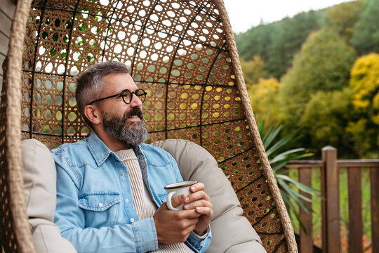 Man is enjoying cup of warm tea, coffee, having relaxing moment at home, sitting in hanging chair on patio.