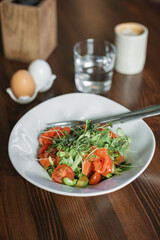 Bowl with tasty salad consisting of lightly salted salmon, arugula, cherry tomatoes, green beans, microgreens, avocado, seasoned with olive oil and lemon juice on wooden table.