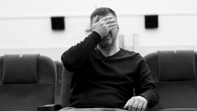 A handsome middle-aged man sits in an armchair in the hall of the village theater, looks at the stage, covers his face with his palm and turns his head disapprovingly, black white video