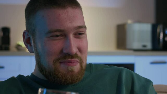 Medium closeup of Caucasian man with beard enjoying his glass of beer and laughing while watching hilarious TV program at night in his modern apartment