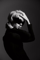 Stylish woman with short blonde hair in black outfit and sunglasses, posing dramatically against a dark grey background. Fashion and confidence concept.