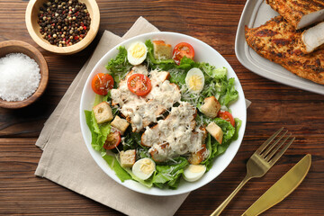 Tasty Caesar salad with chicken and tomatoes in bowl served on wooden table, flat lay