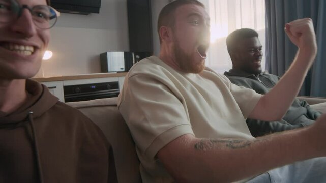 Zoom-in shot of three cheerful young diverse men with sports-themed props in hands getting excited while their favorite team scoring goal or winning, watching game on TV in apartment