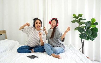 Young asian teen and her friend with headphones listening to music while sitting in the bed to relax in the bedroom, enjoy leisure weekend at home. Stress free concept