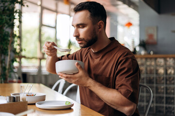 Portrait of handsome bearded man eating Chinese dish holding plate and spoon, sitting in modern cafe