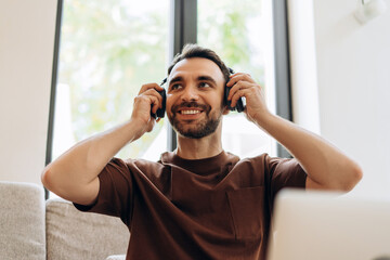 Handsome, smiling bearded man wearing earphones, listening music, looking away, sitting on sofa