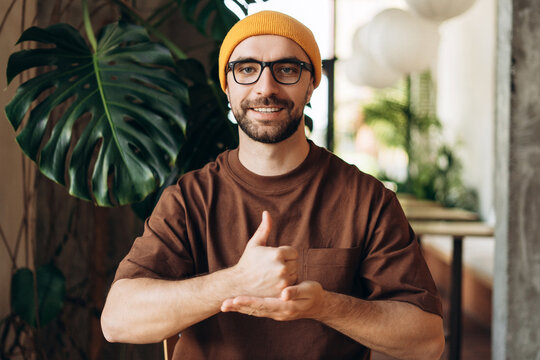 Smiling happy Latin man speaking with sign language, showing thumb up, wearing yellow hat eyeglasses