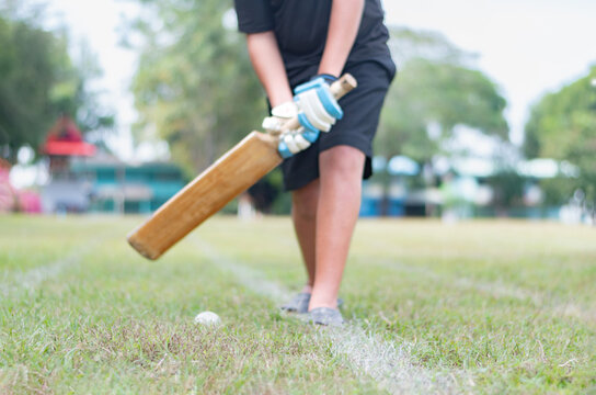 Asian child wears gloves, holding cricket bat and testing to hit cricket ball on outdoor field of cricket sport, soft focus.
