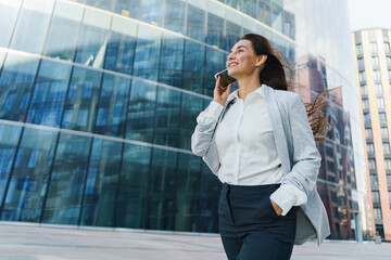 Businesswoman walks confidently while talking on her smartphone near modern glass buildings in an...