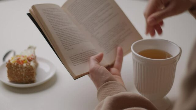 Close-up of a hand holding an open book above a cozy table setup with a cup of tea and a slice of pie. Perfect for relaxation, reading, or lifestyle content