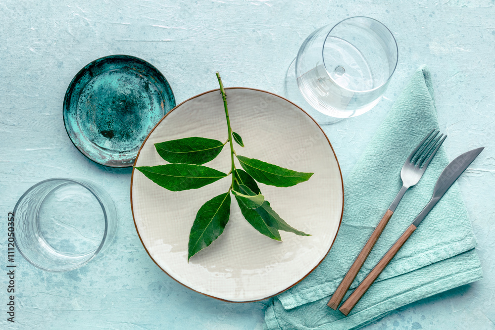 Wall mural Modern tableware, overhead flat lay shot. A table setting with a plate, cutlery, and glasses, shot from the top with green leaves