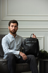 A man is comfortably sitting in a chair, resting with a leather backpack near him