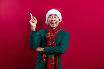 An Asian man in a Santa hat, green sweater, and plaid scarf points upward with an excited expression against a vibrant red background, showcasing holiday joy and enthusiasm in a festive setting