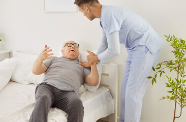 Patient young male caregiver trying to help indignant elderly man to settle into bed. Angry...