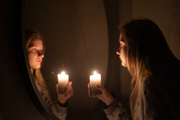 A young woman holds a lit candle, gazing at her reflection in a mirror. The warm glow of the candle...