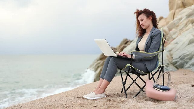 Woman in office clothes, in suit, sitting on chair and working on laptop on seashore. High quality 4k footage