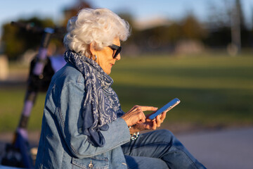 white-haired elderly woman wearing denim sitting in suburban park and swiping on her phone