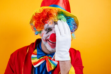 Playful clown hides face, bright yellow background.