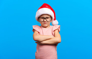 Obraz premium Angry little girl wearing a Santa hat crosses her arms in defiance. Her frustrated expression contrasts with the bright blue background, creating a dramatic holiday scene...