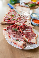 Two florentine steaks seasoned with rosemary and salt, ready to be cooked, resting on a white plate on a wooden table