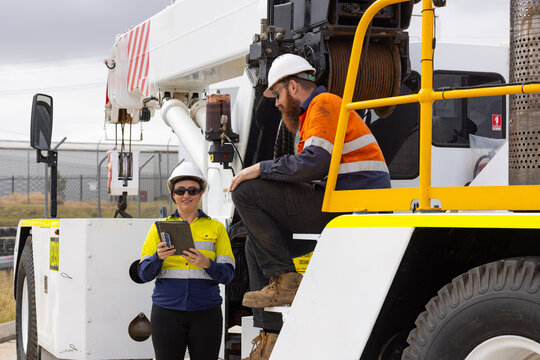 Two workers wearing high visibility clothing man sitting on crane while woman shows him a tablet